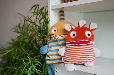 Zwei bunte Sorgenfresser-Kuscheltiere sitzen auf einem weißen Regalbrett vor einer Grünpflanze.