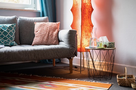 Einblick in einen Beratungsraum mit einem grauen Sofa, einer orangene Lampe und einem Beistelltisch mit Taschentüchtern und einer Wasserkaraffe. 