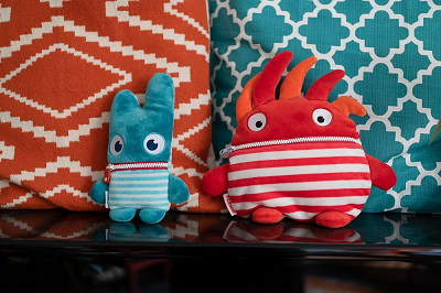 Ein kleines blaues Sorgenfresser-Kuscheltier und ein größeres orangenes Sorgenfresser-Kuscheltier stehen auf einem Regal vor einem orangenen und einem blauen Kissen.